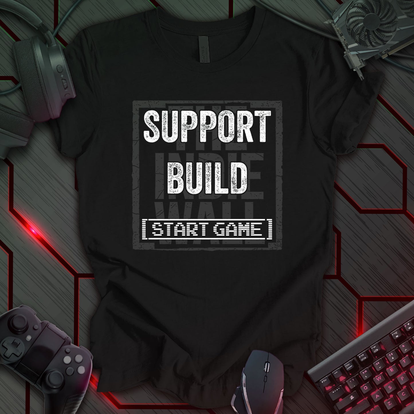 Black gaming t-shirt featuring a distressed typographic design over The Indie Wall logo. The bold white text reads "SUPPORT" and "BUILD," with a retro computer screen-style font below that reads "START GAME."