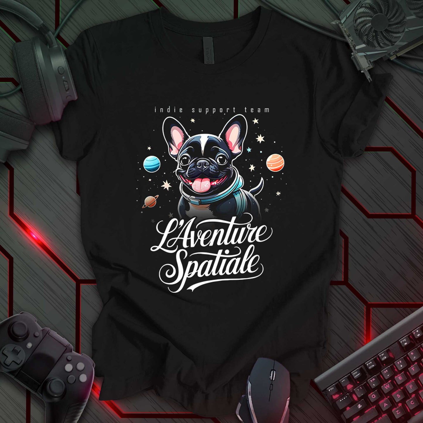 Black t-shirt featuring a whimsical design of a French Bulldog puppy wearing a space suit collar, set against a cosmic backdrop with stars and colorful planets. Elegant script text below reads "L'Aventure Spatiale," while clean text above says "indie support team," making it a fun space-themed gift for gamers and pet lovers.