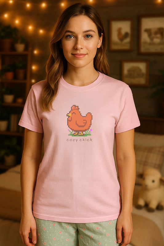 young woman wearing a light pink t-shirt with a chest print of a pixel art image of a cute brown chick standing on a patch of grass. text beneath the pixel art reads "cozy chick". background is an out of focus cozy themed bedroom.