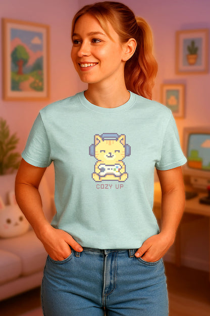 Young woman wearing a light blue t-shirt with a chest print of a pixel art orange cat wearing headphones and holding a game controller above the words "Cozy Up" in cozy gamer kawaii style. the background is a cozy gamer room just out of focus. 