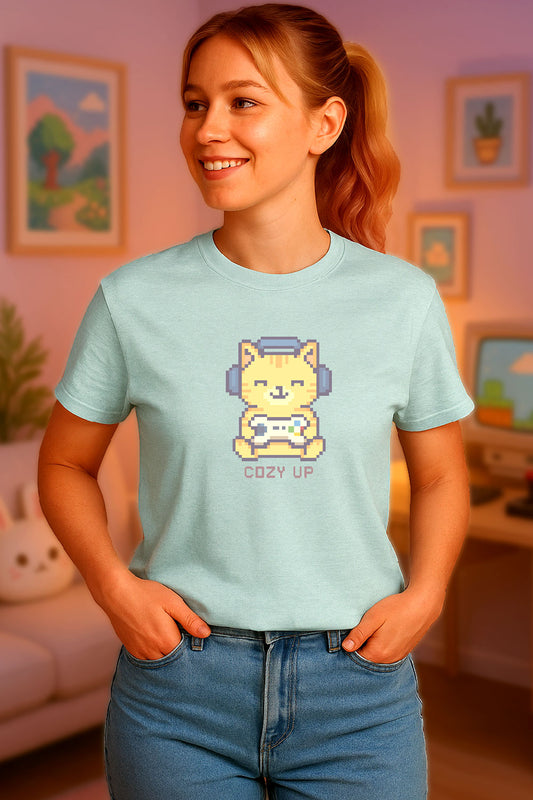Young woman wearing a light blue t-shirt with a chest print of a pixel art orange cat wearing headphones and holding a game controller above the words "Cozy Up" in cozy gamer kawaii style. the background is a cozy gamer room just out of focus. 