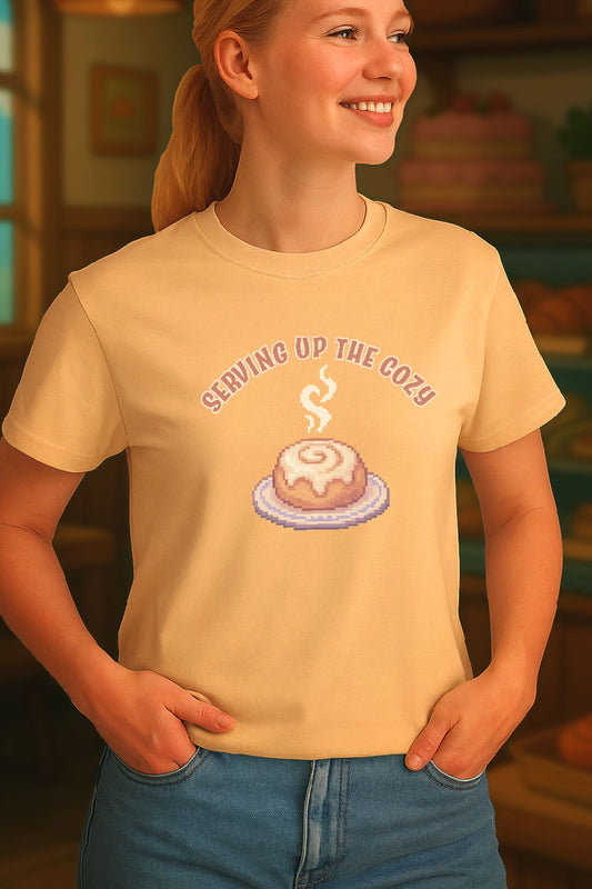 Young woman standing in front of a cozy themed illustration of bakery. she is wearing a t-shirt with a design of pixel art cinnamon roll with steam on a plate under text “Serving Up the Cozy” in cozy gamer kawaii style. The print is on the front of the shirt across the chest. 