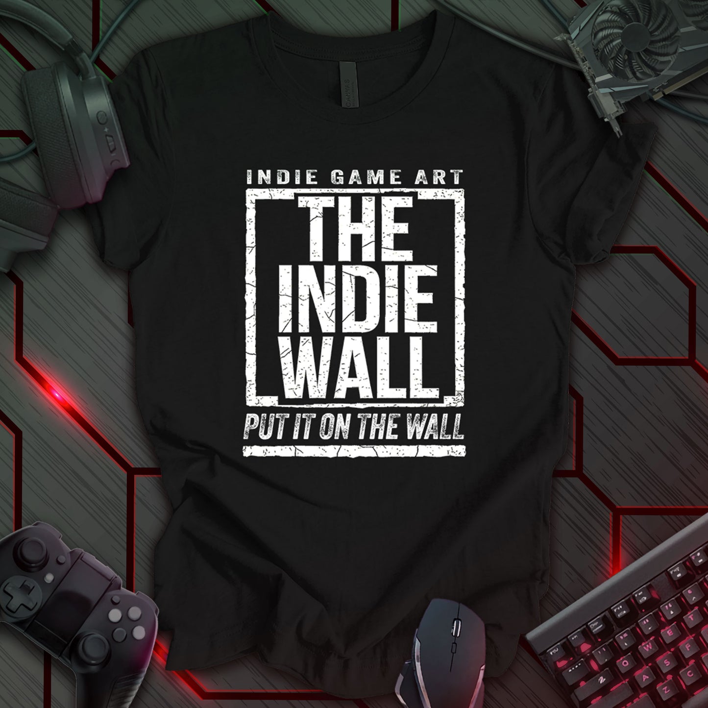 Black gaming t-shirt featuring The Indie Wall logo in distressed bold white text. The design includes "INDIE GAME ART" above the logo and "PUT IT ON THE WALL" below, framed within a textured rectangular border.