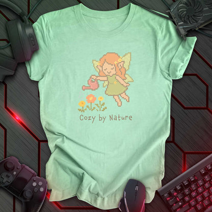 Pixel art fairy with leafy wings watering flowers using a pink watering can, above the text "Cozy by Nature" in cozy kawaii gamer style. Heather Mint background.