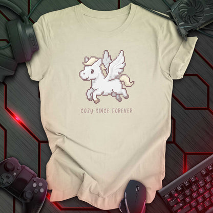 natural color t-shirt with a chest print of a pixel art cute winged Pegasus flying about handwritten styled  text that reads "COZY SINCE FOREVER". 