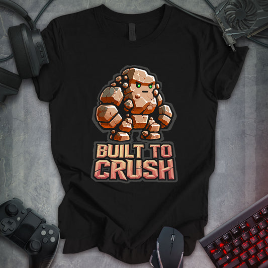 Black gaming t-shirt featuring a cartoon-style rock monster composed of chunky boulder shapes with large arms, legs, and bright green eyes. Below the rock monster, bold stone-text reads 'BUILT TO CRUSH,' designed in a rugged, carved stone font.