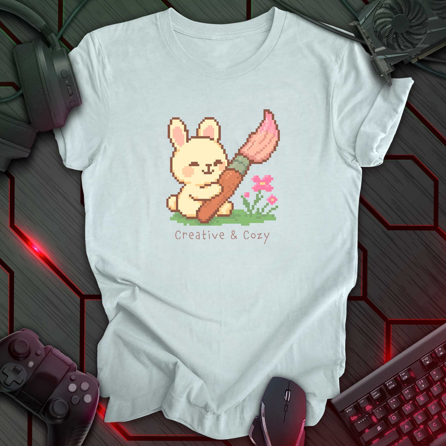 Cute pixel art bunny holding a big pink paintbrush with flowers and grass, under the text "Creative & Cozy" in kawaii style. Heather Ice Blue t-shirt.