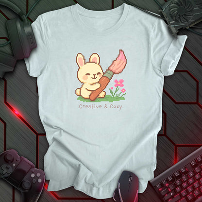 Cute pixel art bunny holding a big pink paintbrush with flowers and grass, under the text "Creative & Cozy" in kawaii style. Heather Ice Blue t-shirt.