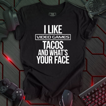 Graphic design featuring the phrase 'I LIKE' in a bold, slightly distressed font at the top, followed by 'VIDEO GAMES' with a rectangle boarder, 'TACOS', and 'AND WHAT'S YOUR FACE' each on separate lines in the same bold, distressed font. All text is white against a heather black background. This is a humorous design with an indie gaming and food theme, featuring a playful and quirky sentiment.
