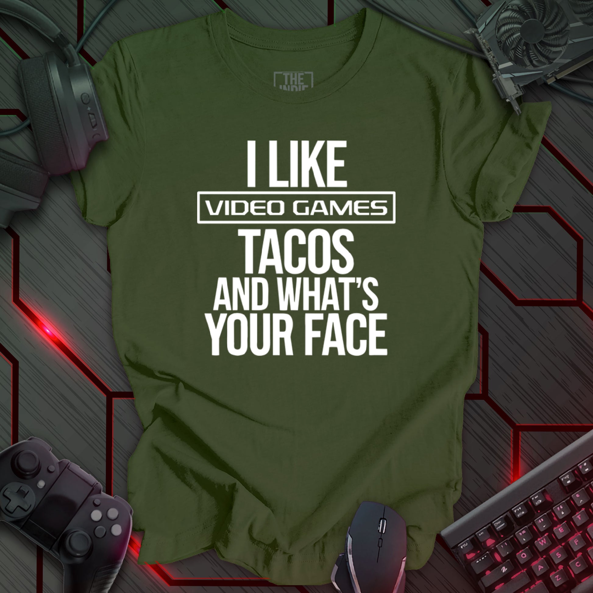 Graphic design featuring the phrase 'I LIKE' in a bold, slightly distressed font at the top, followed by 'VIDEO GAMES' with a rectangle boarder, 'TACOS', and 'AND WHAT'S YOUR FACE' each on separate lines in the same bold, distressed font. All text is white against a military green background. This is a humorous design with an indie gaming and food theme, featuring a playful and quirky sentiment.
