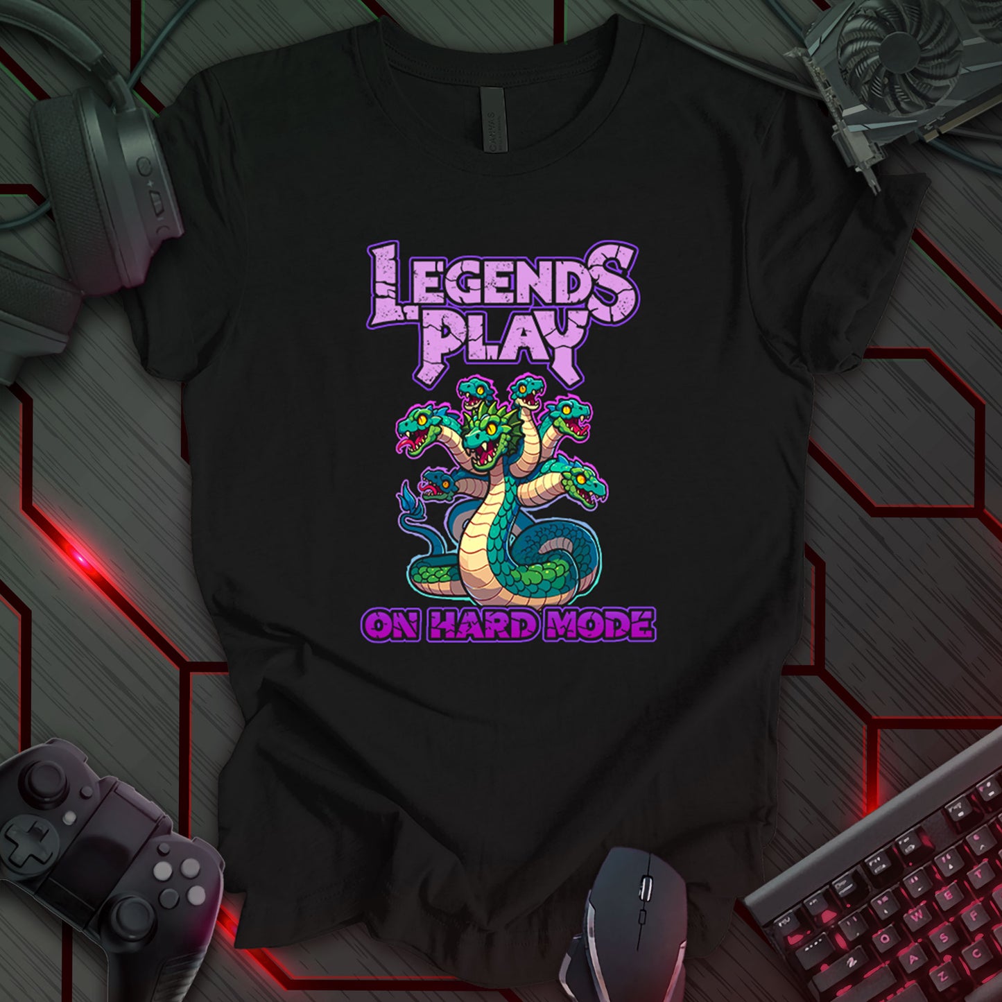 Black gaming t-shirt featuring a cartoon-style seven-headed hydra with green and blue scales, bright yellow eyes, and a bold purple outline. Above and below the hydra, bold stone-themed text in pinkish-purple reads 'Legends Play on Hard Mode.'