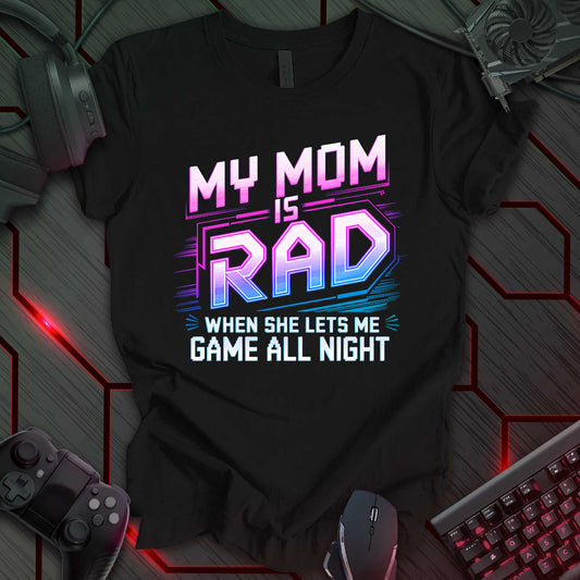 This t-shirt design, targeted at young gamers, features the phrase 'MY MOM IS RAD WHEN SHE LETS ME GAME ALL NIGHT' in a bold, stylized font with pink, blue, and white colors. The design includes geometric lines and shapes, appealing to a playful, video game-loving audience and highlighting the relatable experience of gaming with parental support.