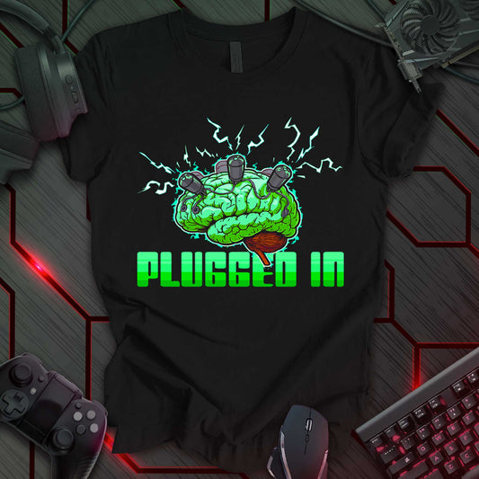 Digital illustration of a green brain with a textured surface, centered against a black background. Three black plugs with wires are inserted into the top of the brain. A brown brain stem is visible at the bottom. Green lightning bolt shapes radiate from the brain. Below the brain is the phrase 'PLUGGED IN' in a blocky, outlined font, rendered in gray. This is a design with a technology or cyberpunk theme, featuring a brain with plugs and the phrase 'PLUGGED IN'.