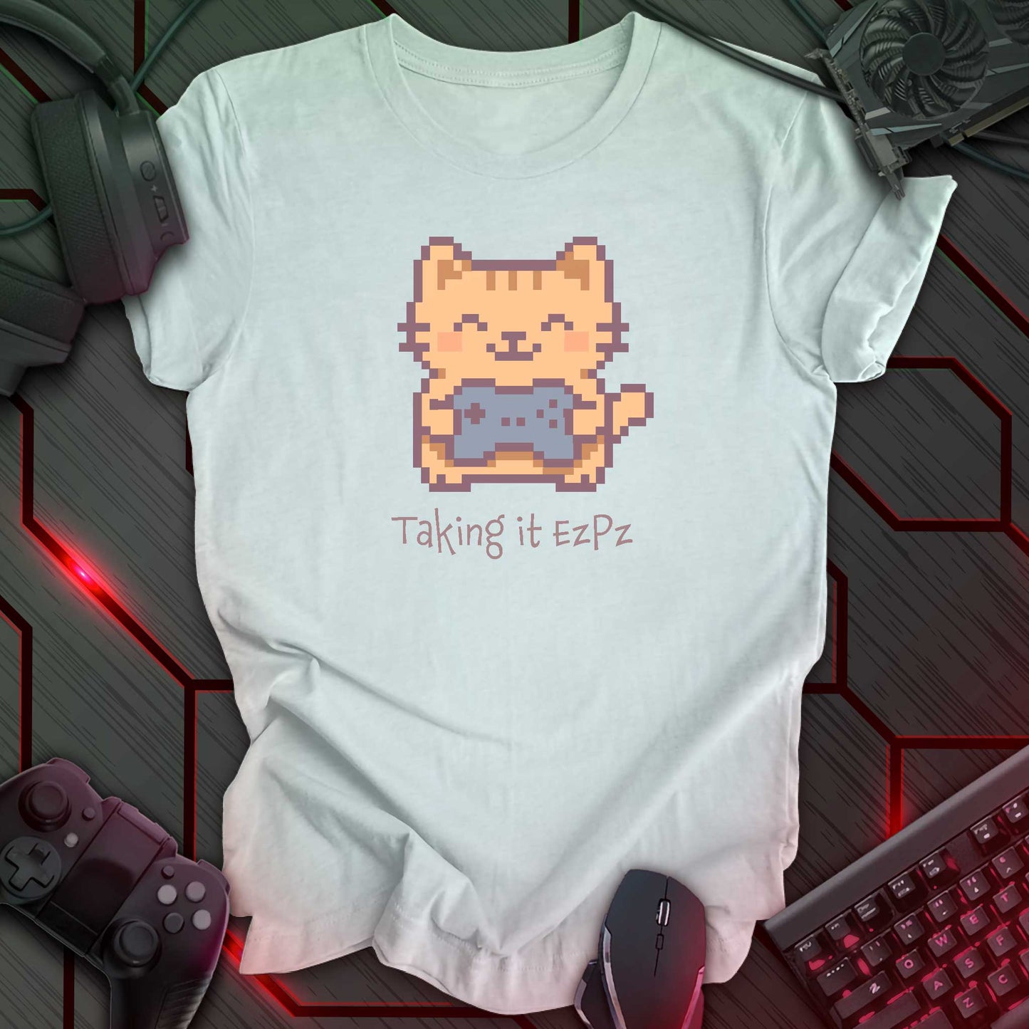 Pixel art cat holding a game controller with closed eyes, smiling above the phrase "Taking it EzPz" in kawaii cozy gamer style. Heather Ice Blue t-shirt.