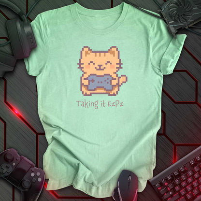 Pixel art cat holding a game controller with closed eyes, smiling above the phrase "Taking it EzPz" in kawaii cozy gamer style. Heather Mint t-shirt.