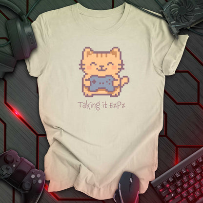 Pixel art cat holding a game controller with closed eyes, smiling above the phrase "Taking it EzPz" in kawaii cozy gamer style. Natural color t-shirt.