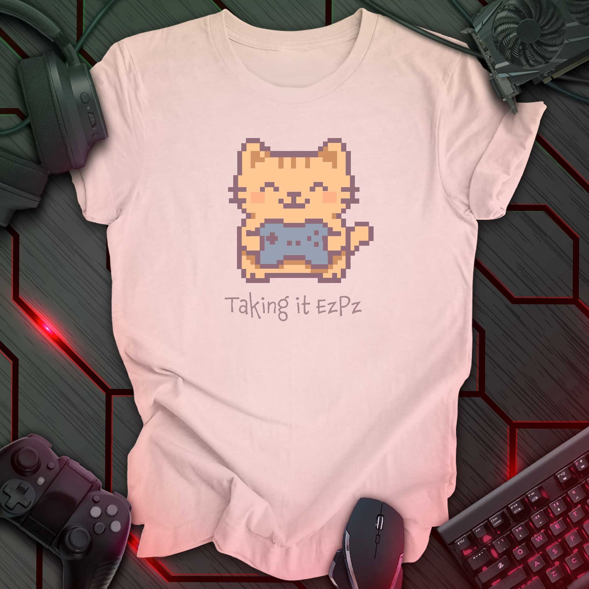 Pixel art cat holding a game controller with closed eyes, smiling above the phrase "Taking it EzPz" in kawaii cozy gamer style. Soft Pink t-shirt.