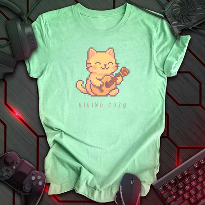 Pixel art cat playing ukulele with cute hand written text “Vibing Cozy” below. Mint green background.